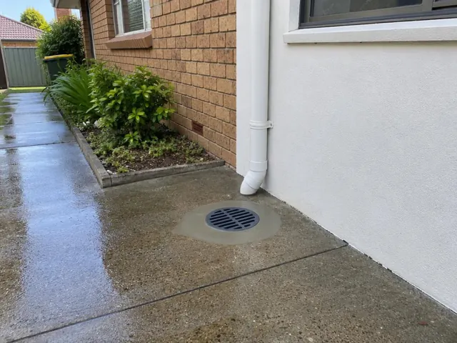 Outdoor drain grate beside a brick house and wet concrete paving in St Marys